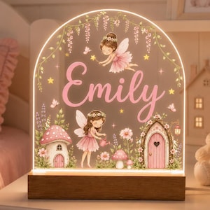 Può includere: Una luce notturna in acrilico ad arco con base in legno che riporta il nome Emily in corsivo rosa. Il design include due fate illustrate con ali, abiti rosa e corone di fiori, circondate da funghi, rampicanti e una piccola porta rosa.