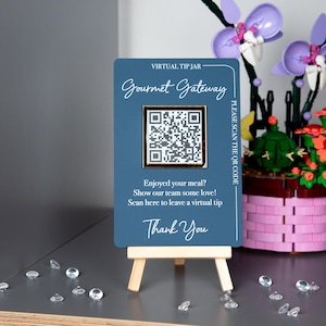 May include: A blue sign on a small wooden easel with the text "Gourmet Gateway" and a QR code. The sign reads "Enjoyed your meal? Show our team some love! Scan here to leave a virtual tip. Thank You."