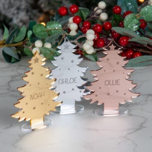 Può includere: Tre segnaposto a forma di albero di Natale in oro, argento e oro rosa. Ogni albero ha un nome inciso: Noah, Chloe e Ollie. Gli alberi sono decorati con stelle e poggiano su basi trasparenti.