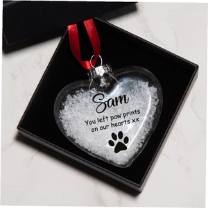 Op de afbeelding: Een glazen hartvormige kerstbal met een sneeuweffect erin. De kerstbal heeft de naam "Sam" en de tekst "You left paw prints on our hearts xx" met een zwarte pootprint.