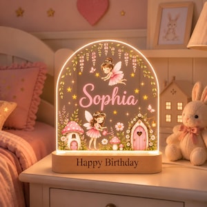 Puede incluir: Una luz nocturna de acrílico en forma de arco con una ilustración de hadas, el nombre Sophia y el texto Happy Birthday. El diseño incluye setas rosas, flores y enredaderas colgantes, iluminadas sobre una base de madera clara.