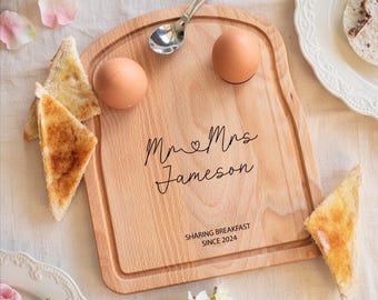 May include: A light-colored wooden breakfast board shaped like a slice of bread, featuring two circular indentations for eggs. The board is engraved with the text "Mr & Mrs Jameson" and "SHARING BREAKFAST SINCE 2024" in a black script and sans-serif font.