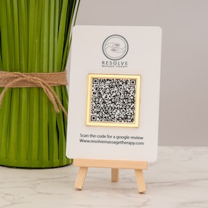 Könnte beinhalten: Eine weiße Karte mit einem goldenen Rahmen um einen schwarz-weißen QR-Code. Die Karte steht auf einem kleinen Holzständer. Auf der Karte steht der Text "RESOLVE MASSAGE THERAPY" und "Scannen Sie den Code für eine Google-Bewertung www.resolvemassagetherapy.com".