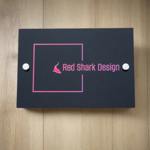 May include: A rectangular black sign with the text "Red Shark Design" in pink, along with a pink shark logo. The sign is mounted with two silver screws on a light wood surface.