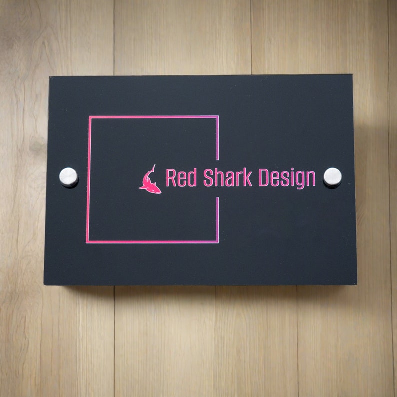 May include: A rectangular black sign with the text "Red Shark Design" in pink, along with a pink shark logo. The sign is mounted with two silver screws on a light wood surface.