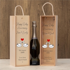 Könnte beinhalten: Zwei Weinflaschenkisten aus Holz mit Seilgriffen, die eine Champagnerflasche flankieren. Die Kisten sind mit Schwanenmotiven, Herzen und dem Text "Happy Ruby Anniversary" und Namen verziert. Eine Kiste sagt "9th August 2000".