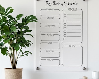 May include: Clear acrylic weekly schedule board with black text. The board includes sections for each day of the week, a to-do list, and notes. The text reads "This Week's Schedule". The board is mounted on a white wall with silver hardware.