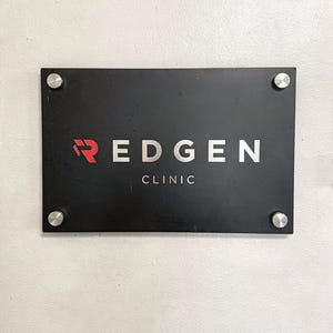 May include: A rectangular black sign with the text "REDGEN CLINIC" in white and red lettering. The sign is mounted on a white wall with four silver screws in each corner.