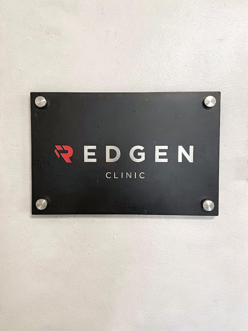 May include: A rectangular black sign with the text "REDGEN CLINIC" in white and red lettering. The sign is mounted on a white wall with four silver screws in each corner.