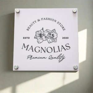 May include: A square, light pink sign with the text "BEAUTY & FASHION STORE" and "MAGNOLIAS" in black. The sign also features a floral illustration and the words "ESTD 2020" and "Premium Quality". The sign is mounted with four silver fasteners.