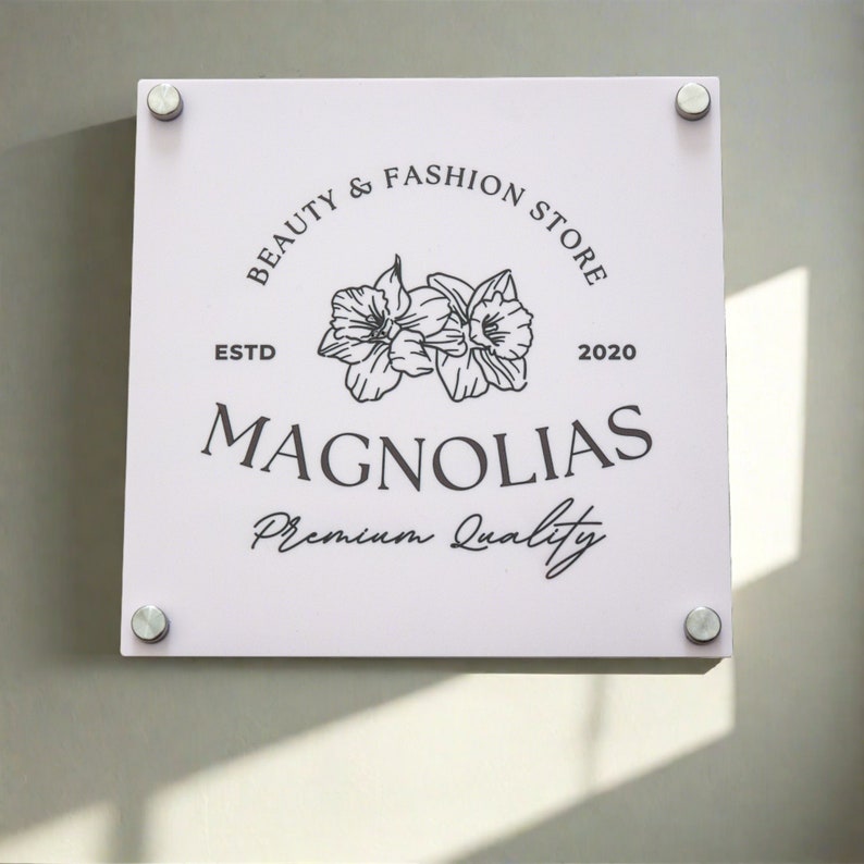 May include: A square, light pink sign with the text "BEAUTY & FASHION STORE" and "MAGNOLIAS" in black. The sign also features a floral illustration and the words "ESTD 2020" and "Premium Quality". The sign is mounted with four silver fasteners.