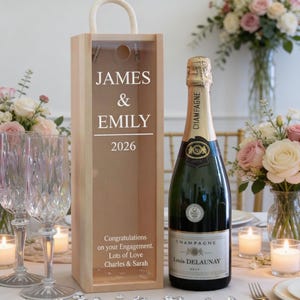 May include: A wooden champagne bottle gift box with the names "JAMES & EMILY" and the year "2026" printed on it. A bottle of champagne and two crystal glasses are on a table with flowers and candles. The box reads "Congratulations on your Engagement. Lots of Love Charles & Sarah."