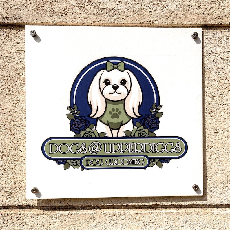 May include: A white square sign with the text "DOGS @ UPPERDIGGS DOG GROOMING". The sign features a cartoon dog with a green paw print on its chest, surrounded by blue roses and a blue and white circular design.