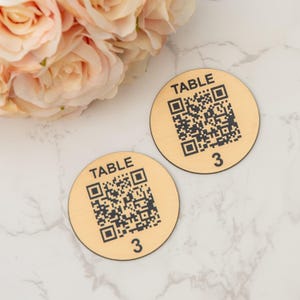 Op de afbeelding: Twee ronde houten tafelnummerborden met een zwart-witte QR-code en de tekst "TABLE 3" erop gedrukt.