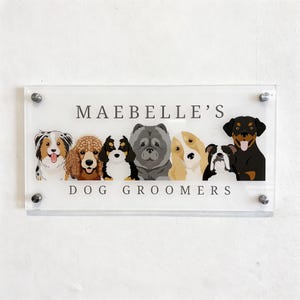 May include: Clear acrylic sign with the text "MAEBBELLE'S DOG GROOMERS" and illustrations of various dog breeds. The sign is mounted with silver hardware and features a white background.