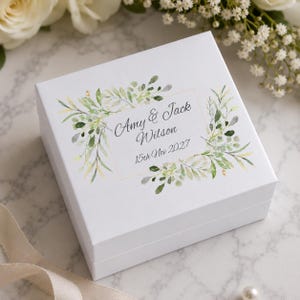 May include: A square white gift box featuring a printed design of green watercolor leaves framing the text Amy & Jack Wilson and 15th Nov 2027. The box has a clean, smooth finish and is positioned at an angle to display the top lid graphic clearly.