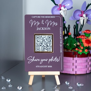 Può includere: Un cartello nuziale color prugna su un piccolo cavalletto di legno. Il cartello recita "Mr & Mrs Jackson" e include un codice QR. Viene visualizzato anche il testo "Share your photos! 6th August 2026". Il cartello è bordato d'oro.