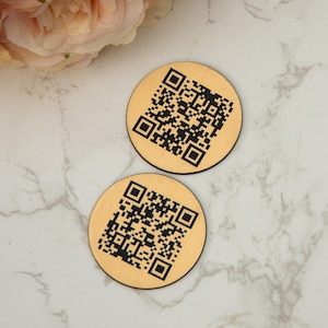 Può includere: Due sottobicchieri rotondi in legno con codici QR in bianco e nero stampati su di essi. I sottobicchieri sono su una superficie di marmo bianco.