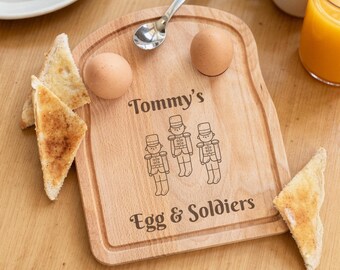 May include: A light-colored wooden serving board shaped like a slice of bread, featuring two circular indentations for eggs, a silver spoon, and an engraving that reads Tommy's Egg & Soldiers with three illustrated toy soldier figures.