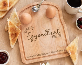 May include: A light-colored wooden serving board shaped like a slice of bread, engraved with the text "DAD's Eggcellent EGGS" and "Happy Birthday Dad, Love Alex & Emma x". It holds two brown eggs and a silver spoon, surrounded by toast.