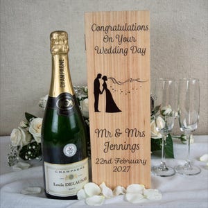 May include: A champagne bottle with a gold foil neck and a label that reads "Champagne Louis Delaunay". Beside it is a wooden box engraved with "Congratulations On Your Wedding Day", a silhouette of a couple, and the names "Mr & Mrs Jennings" and the date "22nd February 2027". Two champagne flutes are also present.