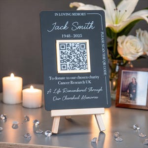Könnte beinhalten: Ein dunkles, graues Gedenkschild auf einer kleinen Holzstaffelei. Auf dem Schild steht "IN LOVING MEMORY" und "Jack Smith 1948-2025". Es enthält einen QR-Code und den Text "PLEASE SCAN THE QR CODE". Auf dem Schild steht auch "To donate to our chosen charity Cancer Research UK A Life Remembered Through Our Cherished Memories."