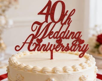 May include: A white iced cake with a red glitter cake topper that reads "40th Wedding Anniversary." The cake is decorated with red heart accents and a pearl border. The topper is in a cursive font.