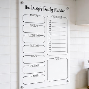 May include: A clear acrylic family planner with the text "The Laing's Family Planner" in black script. It features sections for each day of the week, a to-do list, and notes. The planner is mounted on a white tiled wall with silver hardware.