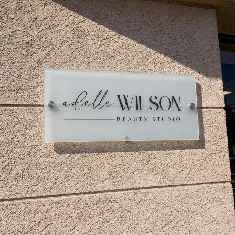 May include: A rectangular glass sign with the text "adelle WILSON BEAUTY STUDIO" in black lettering. The sign is mounted on a textured beige wall with silver hardware.