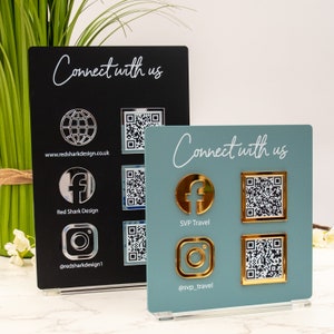 Può includere: Due cartelli in acrilico blu-verde e nero con il testo "Connect with us" in corsivo bianco. Ogni cartello presenta un diverso set di icone di social media e codici QR. Il cartello nero ha un globo argentato, icone di Facebook e Instagram. Il cartello blu-verde ha icone di Facebook e Instagram dorate.