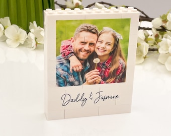 Pode incluir: Uma moldura de foto branca em bloco com uma foto de um pai e sua filha. A foto é um quebra-cabeça, e o texto "Daddy & Jasmine" está escrito em itálico na moldura. Flores brancas e vegetação no fundo.