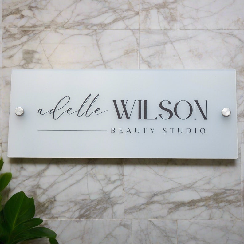 May include: A rectangular frosted glass sign with the text "adelle WILSON BEAUTY STUDIO" in black and gray lettering. The sign is mounted on a marble tile wall with two silver-colored fasteners.