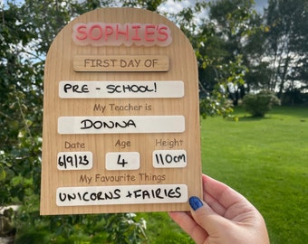 May include: Wooden sign with the text "SOPHIE'S FIRST DAY OF PRE-SCHOOL!" and details including the teacher's name, date, age, height (110cm), and favorite things: UNICORNS + FAIRIES. The sign is held outdoors.
