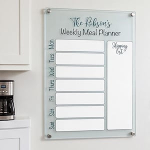 May include: A clear glass weekly meal planner with the text "The Robson's Weekly Meal Planner" and "Shopping List". The days of the week are listed vertically on the left side. The planner is mounted on a white wall with silver fixings.