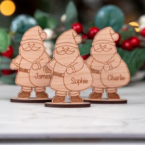 Puede incluir: Tres figuras de madera de Papá Noel con nombres personalizados en sus bolsas de regalo. Cada Papá Noel tiene una base redonda y está hecho de madera clara. Los nombres son "James", "Sophie" y "Charlie". Decoración festiva navideña.