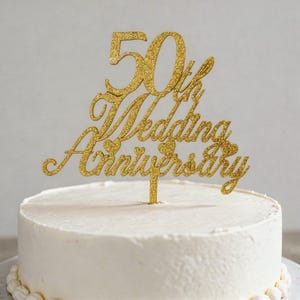 May include: A gold glitter cake topper that reads "50th Wedding Anniversary" is placed on top of a white frosted cake. The topper has a script font with hearts. The cake is on a white surface.
