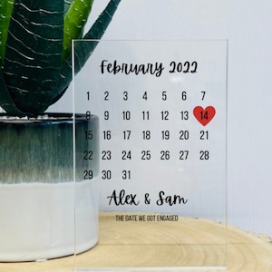 May include: A clear acrylic calendar showing the month of February 2022. The 14th is highlighted with a red heart. The text "Alex & Sam" and "THE DATE WE GOT ENGAGED" is printed below the calendar.