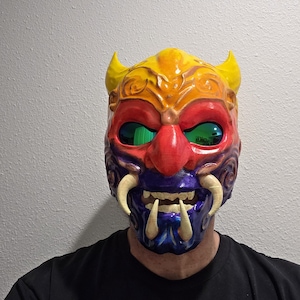 Puede incluir: Una máscara Oni detallada y colorida con cuernos amarillos, ojos rojos y una nariz roja. La máscara presenta una boca morada y azul con grandes colmillos blancos. Es un artículo de cosplay o disfraz.