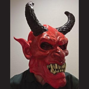 Puede incluir: Una máscara de diablo roja con cuernos negros grandes, orejas puntiagudas y una sonrisa amenazante. La máscara tiene una textura detallada y la lleva una persona. Es un disfraz de Halloween o un accesorio teatral.