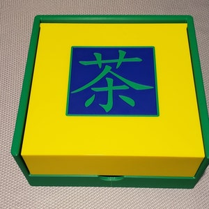 Könnte beinhalten: Eine grüne und gelbe Box mit einem blauen Quadrat in der Mitte. Das Quadrat enthält das chinesische Zeichen für "Tee" in grün.