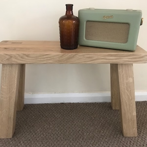 Puede incluir: Un banco de madera rústico con un acabado natural y cuatro patas. Una botella de vidrio marrón y una radio vintage verde están encima del banco.