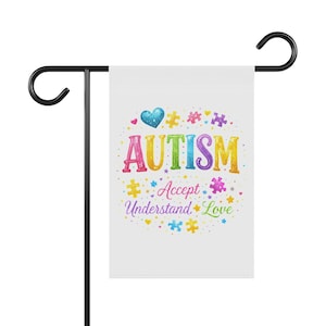 May include: A white garden flag with the word "AUTISM" in colorful letters, along with the words "Accept, Understand, Love." The flag has a blue heart and puzzle pieces. The flag is attached to a black metal garden stake.