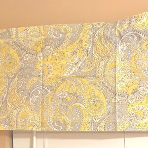Op de afbeelding: Een decoratieve valance met een paisley-patroon in tinten geel, grijs en wit. De stof is gevouwen en gedrapeerd, waardoor een textuur ontstaat. De valance wordt waarschijnlijk gebruikt voor raambekleding.