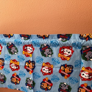 Op de afbeelding: Lichtblauwe stof met kleurrijke cartoonafbeeldingen van Paw Patrol-personages, waaronder Chase, Marshall en Rocky. Het patroon herhaalt de badges van de personages en het Paw Patrol-logo.