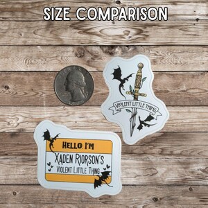 May include: Two stickers with dragon and sword designs, and a quarter for size comparison. One sticker reads "VIOLENT LITTLE THING" with a sword and dragons. The other sticker is a name tag that says "HELLO, I'M XADEN RIORSON'S VIOLENT LITTLE THING".