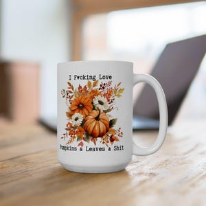 May include: White ceramic mug with a handle, featuring a fall-themed design. The design includes orange pumpkins, white flowers, and autumn leaves. Text on the mug reads "I Fucking Love Pumpkins & Leaves & Shit."