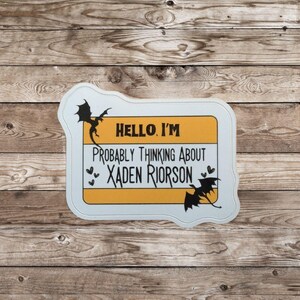 May include: A white and yellow name tag with the text "HELLO, I'M PROBABLY THINKING ABOUT XADEN RIORSON" and dragon illustrations. The name tag is on a wooden background.