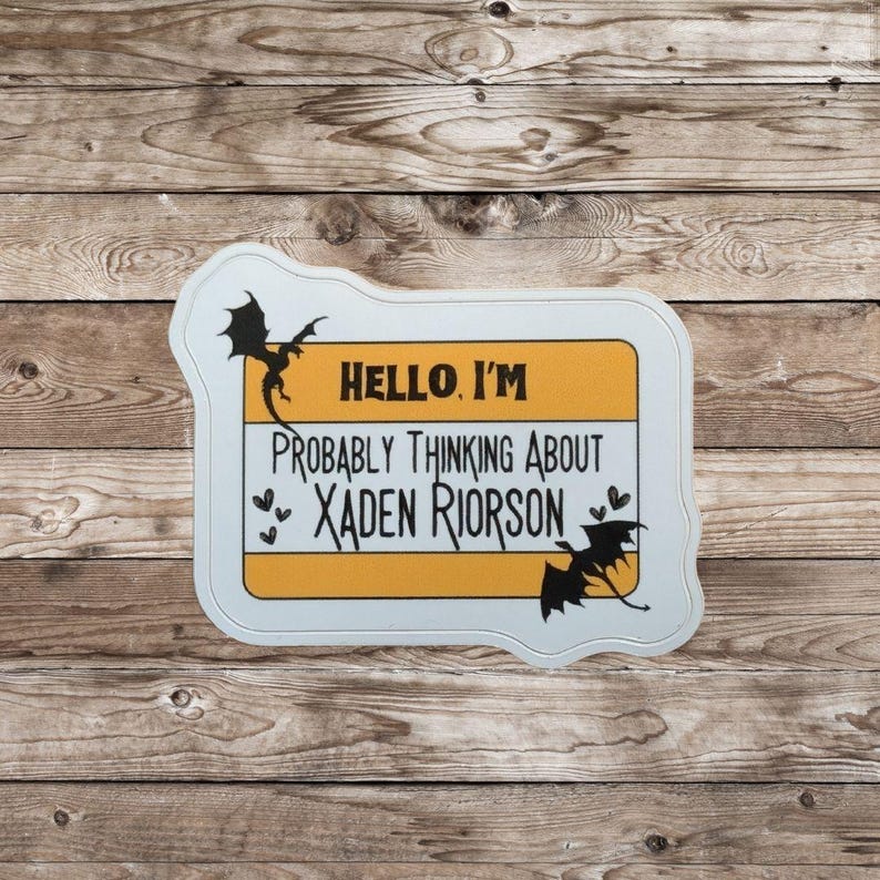 May include: A white and yellow name tag with the text "HELLO, I'M PROBABLY THINKING ABOUT XADEN RIORSON" and dragon illustrations. The name tag is on a wooden background.
