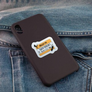 May include: A dark brown phone case with a sticker that reads "Hello I'm Xaden Riorson's Violent Little Thing" in black text on a yellow and white background. The sticker has bat illustrations. The case is on a denim jacket.