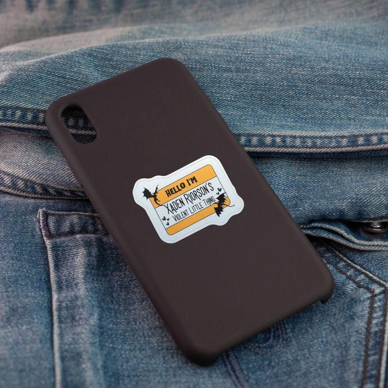May include: A dark brown phone case with a sticker that reads "Hello I'm Xaden Riorson's Violent Little Thing" in black text on a yellow and white background. The sticker has bat illustrations. The case is on a denim jacket.